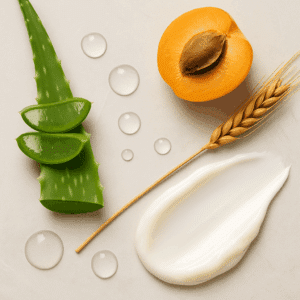 A flat-lay arrangement featuring fresh aloe slices, half an apricot with its pit, a wheat stalk, water droplets, and a swipe of creamy conditioner on a light surface, representing natural hydrating and strengthening ingredients.